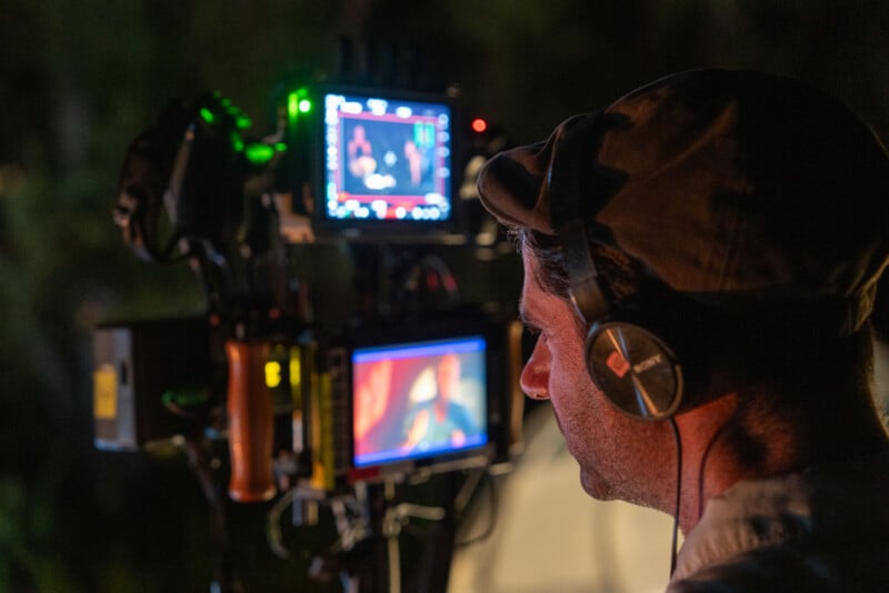 Un hombre con auriculares y sombrero opera una cámara profesional con dos monitores que muestran imágenes en color, posiblemente mientras graba una película o un vídeo por la noche.