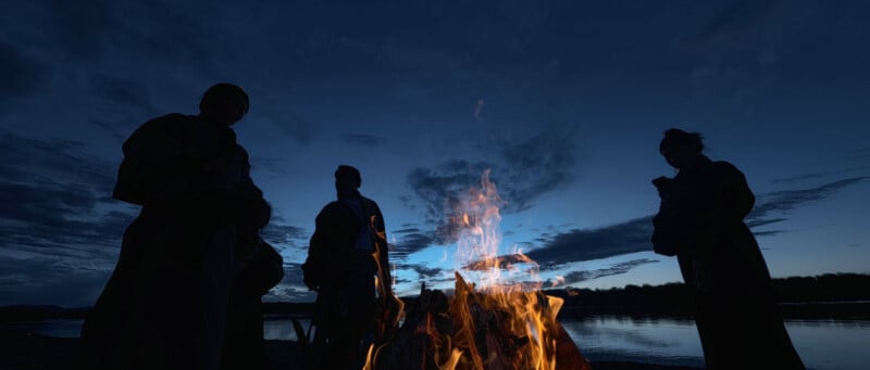 Cuatro figuras vestidas con túnicas se encuentran alrededor de una fogata cerca de una masa de agua al anochecer, con un telón de fondo de espectaculares cielos nublados y un paisaje oscuro.