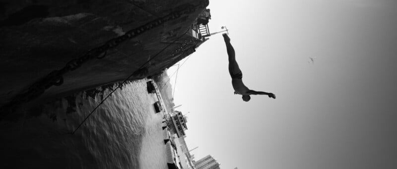 Un hombre en traje de baño salta desde un muelle de concreto al agua; la imagen es en blanco y negro y parece girar, con edificios y un río visibles al fondo.