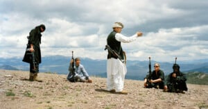 Four people in dark clothing holding rifles sit or stand on rocky ground, while a person in white and black traditional clothing gestures to them. Mountains and a cloudy sky are visible in the background.