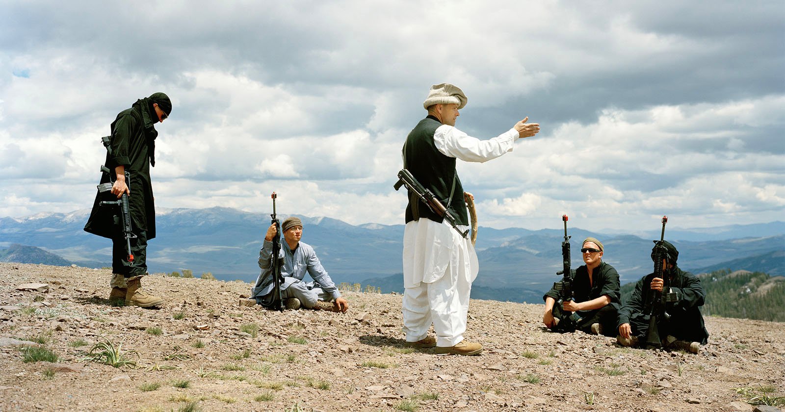 Four people in dark clothing holding rifles sit or stand on rocky ground, while a person in white and black traditional clothing gestures to them. Mountains and a cloudy sky are visible in the background.