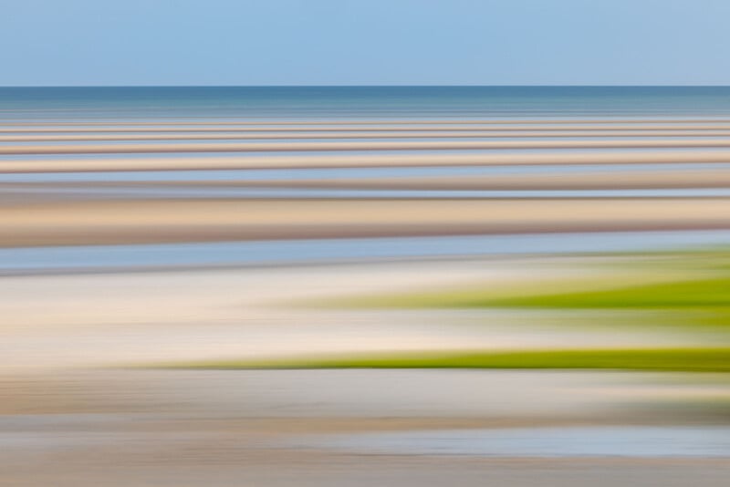 Abstract image of a beach landscape with horizontal stripes in shades of blue, brown, white, and green, suggesting water, sand, and grass, slightly blurred to create a sense of motion.