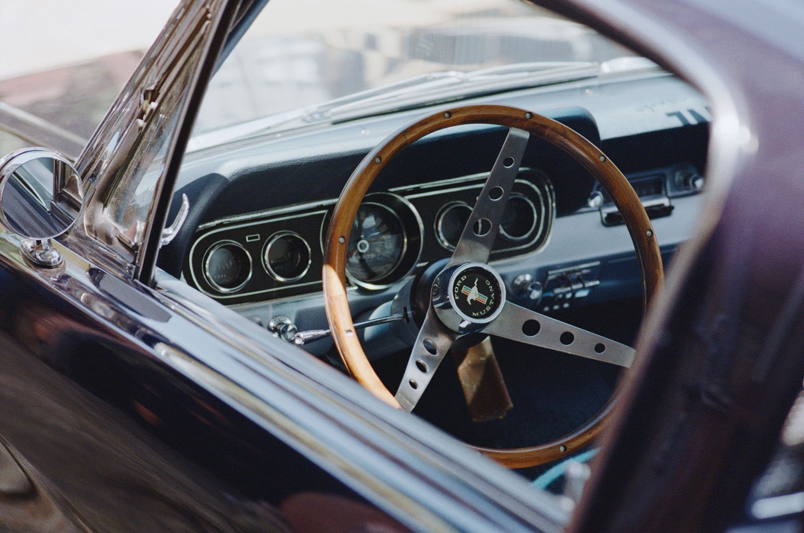 Close-up view of the interior of a classic car, showing a wooden steering wheel with metal spokes, dashboard dials, and part of the driver’s seat through an open door.