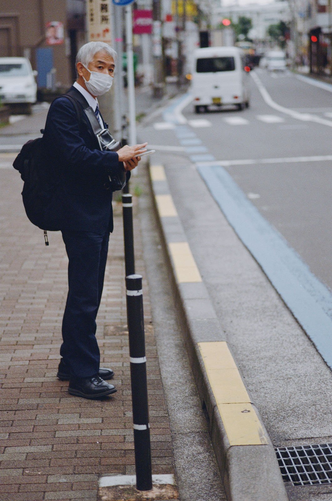 An older man in a suit, wearing a face mask and carrying a backpack, stands on a city sidewalk near the curb, looking at his phone. A white van drives away on the empty street in the background.