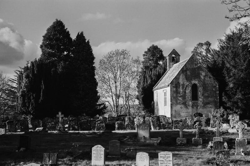 A small, old stone church stands among many weathered gravestones in a cemetery, surrounded by tall trees. The scene is in black and white, with partly cloudy skies above.