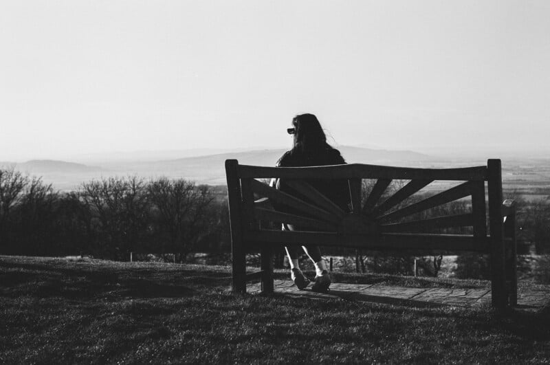 A person sits alone on a wooden bench, facing a scenic landscape with distant hills and trees. The image is in black and white, and the sunlight casts long shadows across the grass.