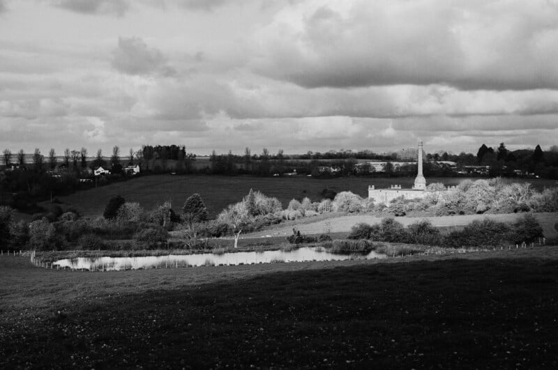 A black and white photo of a rural landscape with open fields, a church with a tall steeple, scattered trees, a pond, and houses in the distance under a cloudy sky.