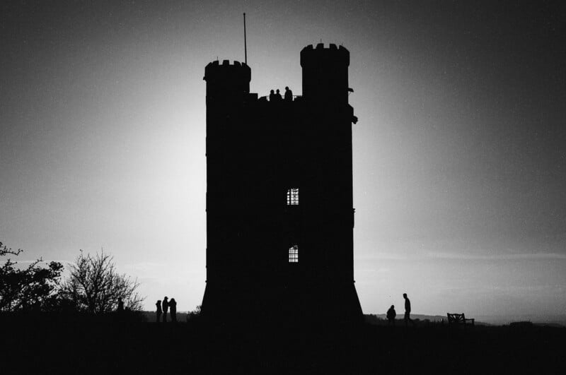 A tall, two-towered castle is silhouetted against the bright sky, with a few people standing and walking nearby, creating striking shadows in the foreground.