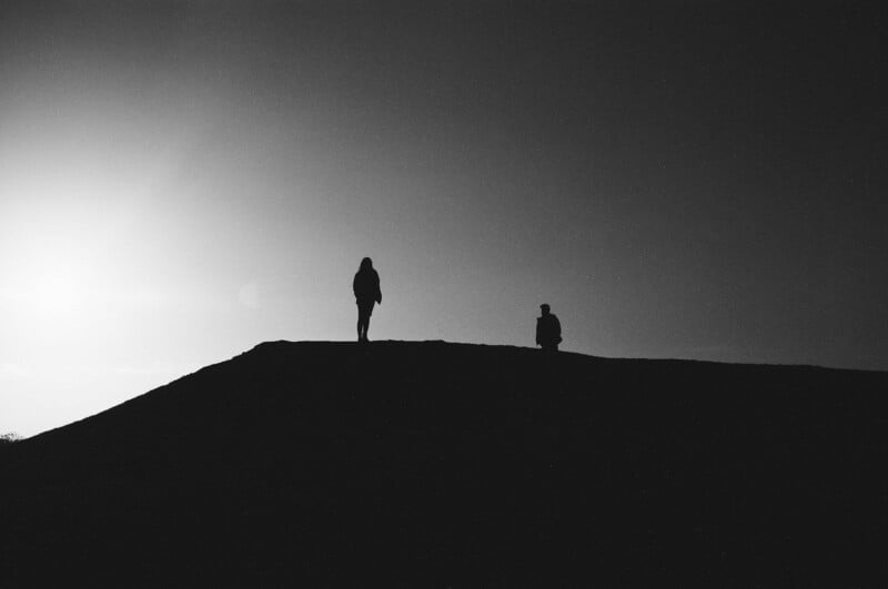 Two people are silhouetted against a bright sky, standing and sitting atop a hill, creating a dramatic contrast between light and shadow.