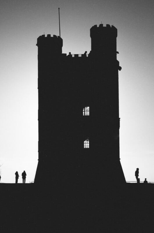 Silhouette of a tall, twin-towered castle with a flagpole on top, backlit by the sky. Four people stand at the base, also in silhouette, while three windows in the castle glow with light.