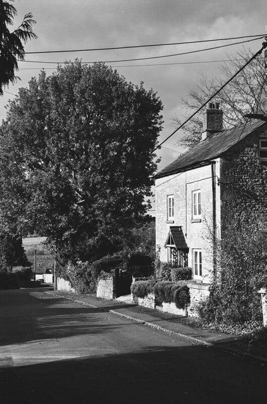 Black and white photo of a quiet village street with a tall tree and a two-story brick house. The house has a porch, chimney, bushes out front, and power lines overhead under a partly cloudy sky.