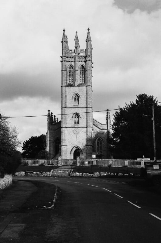 Black and white photo of a stone church with a tall, ornate tower, arched windows, and pointed spires, set behind a low fence and trees, with a curved road in the foreground.