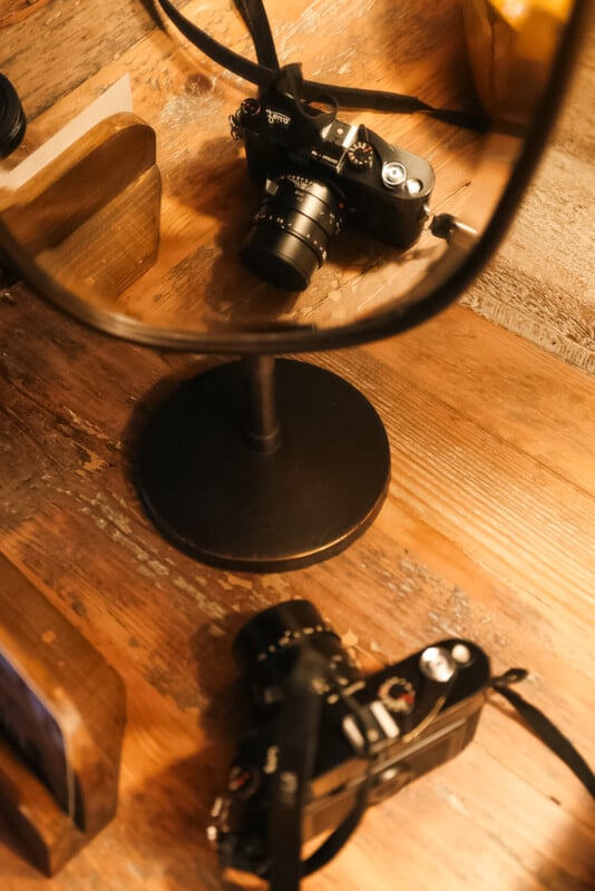 A black camera rests on a wooden surface, reflected in a round mirror with a black metal stand, creating a double image of the camera. Warm lighting highlights the wood grain and camera details.