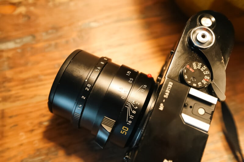 A close-up photo of a black Leica camera with a 50mm lens and visible focus and aperture markings, placed on a wooden surface.