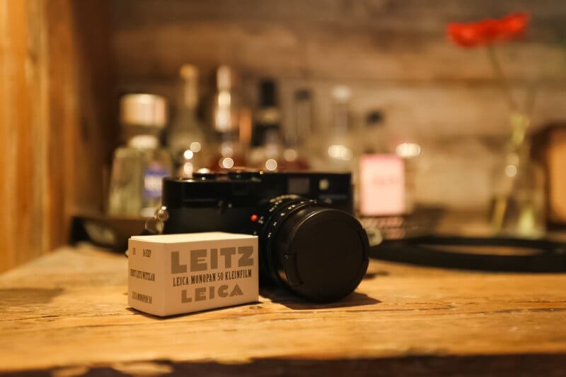A Leica camera with a lens cap rests on a wooden surface next to a box labeled "Leitz Leica," with blurred bottles and a red flower in the background.