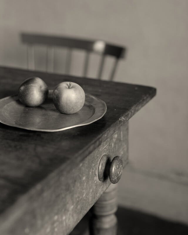 Una fotografía en blanco y negro muestra dos manzanas sobre una placa de metal sobre una mesa rústica de madera, con el respaldo de una silla de madera visible al fondo.