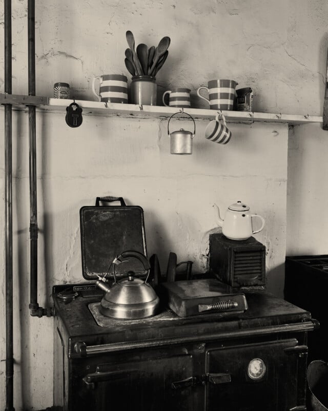 Escena de cocina retro con estufa de metal, hervidor y tetera. En el estante superior hay tazas a rayas, cuencos, una olla con cubiertos y una taza colgante. La imagen está en blanco y negro.