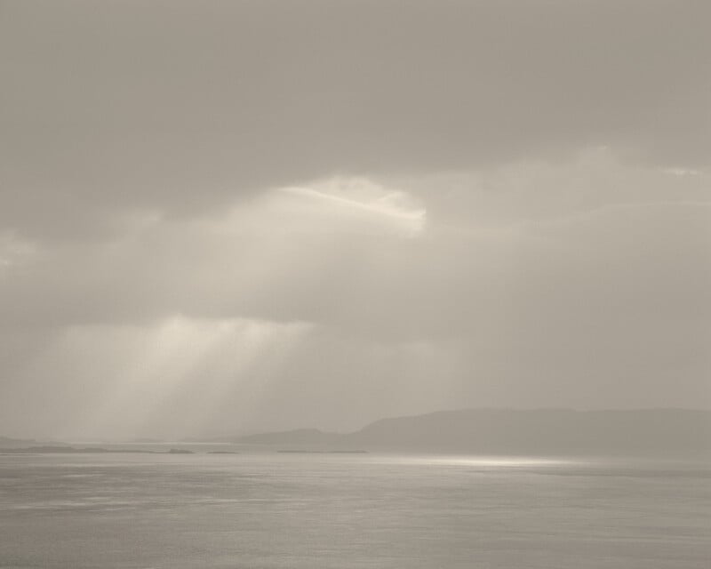 La suave luz del sol atraviesa las espesas nubes, proyectando rayos sobre el agua tranquila y reflectante, con la silueta de montañas distantes al fondo. La escena es serena y monocromática.