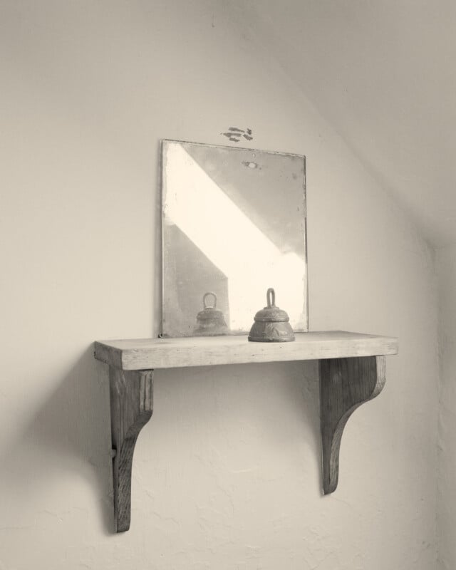 En una habitación suavemente iluminada, dos campanas de metal se encuentran en un pequeño estante de madera en la pared, frente a un viejo espejo cuadrado que refleja la luz, apoyado contra una pared blanca texturizada.