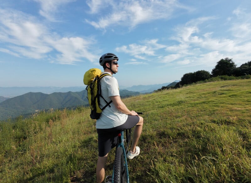 Un hombre que lleva un casco y una mochila amarilla anda en bicicleta sobre una colina cubierta de hierba verde, mirando las montañas distantes bajo el cielo azul y las nubes blancas.