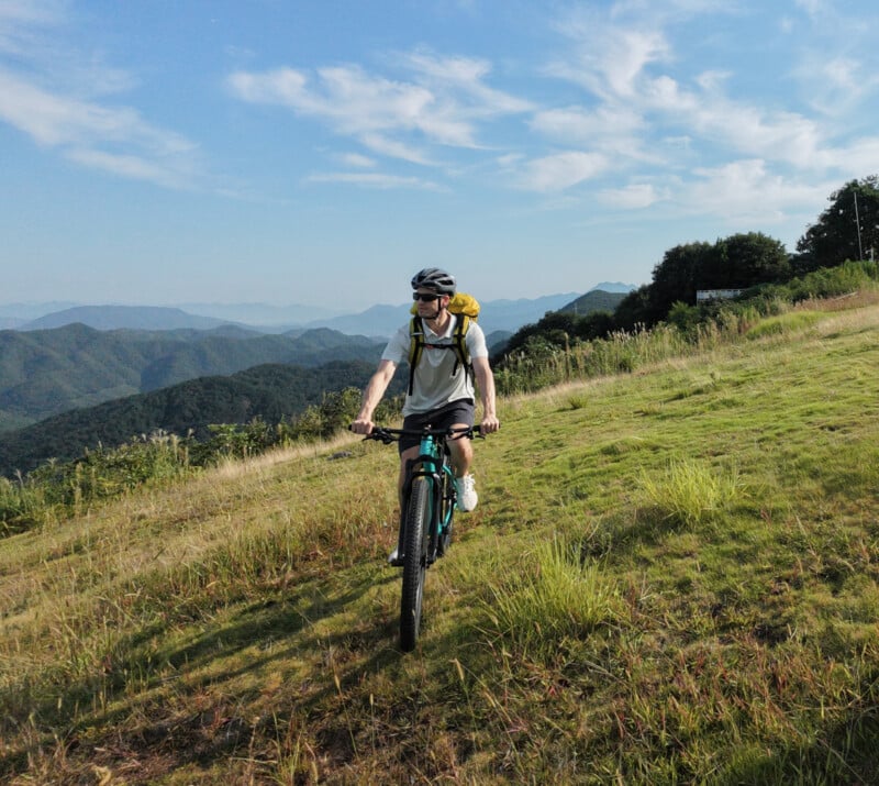 Un hombre que lleva un casco y una mochila monta una bicicleta de montaña cuesta arriba en una pendiente cubierta de hierba, con montañas verdes, agua verde, cielo azul y nubes blancas al fondo.