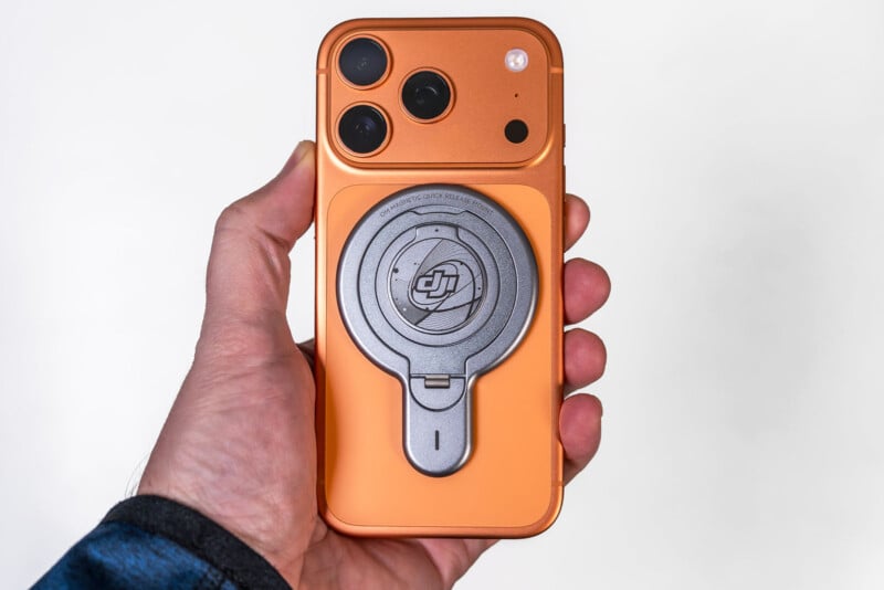 En una mano sostiene un teléfono inteligente naranja con tres cámaras traseras y un accesorio DJI redondo de metal adjunto a la parte posterior. El fondo es de un blanco puro.