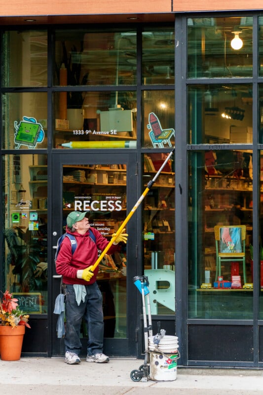 A window cleaner in a red jacket and yellow gloves uses a long pole to clean the glass door of a store called "Recess," with cleaning supplies and plants visible nearby.