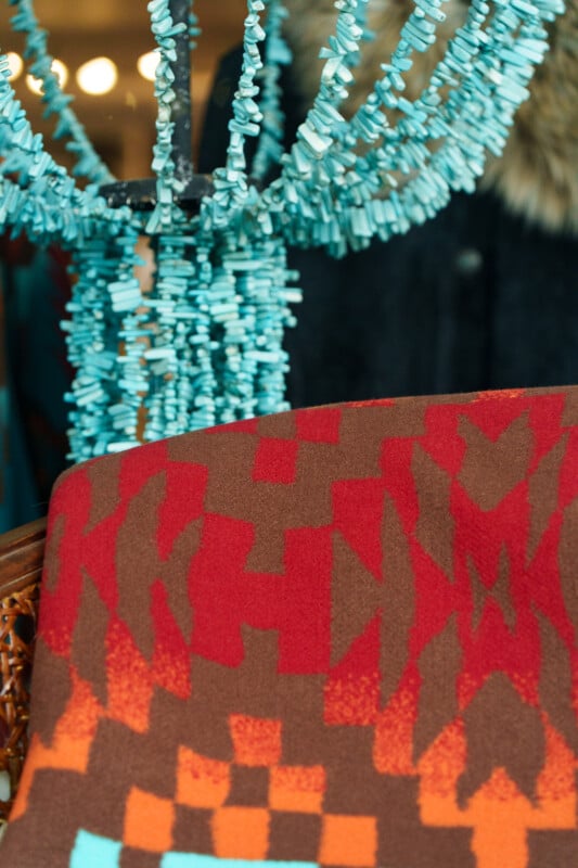 A close-up of a geometric-patterned brown, red, and orange blanket with a turquoise beaded necklace draped on a chair in the background.