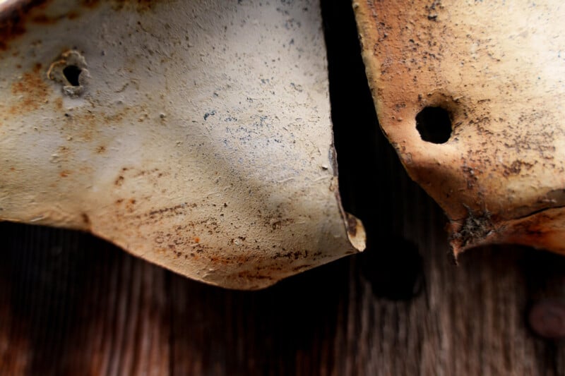 Close-up of two pieces of rusted, weathered metal with holes, set against a wooden surface. The metal shows signs of age and corrosion, creating a textured and rough appearance.