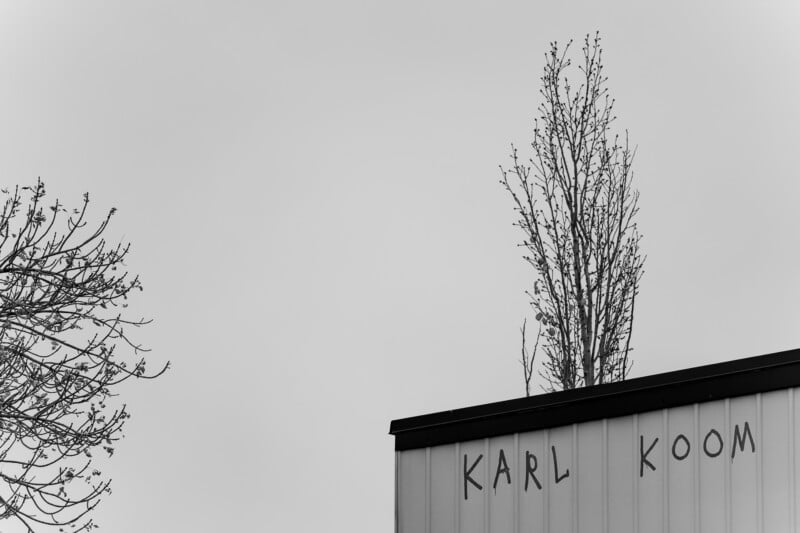 A black-and-white photo shows bare trees and the corner of a building with “KARL KOOM” written on its side against a cloudy sky.
