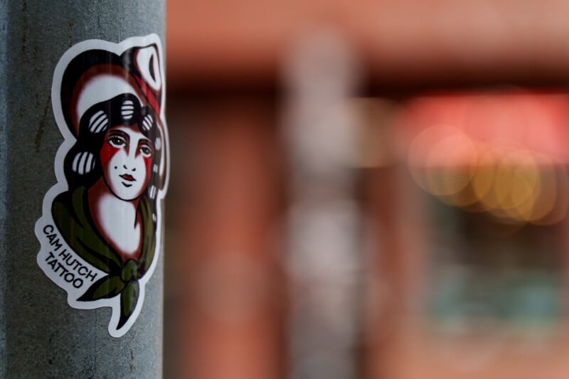 A sticker of a traditional-style tattoo portrait of a woman with a red hat and green scarf is placed on a gray pole. The background is blurred with warm colors and bokeh lights.