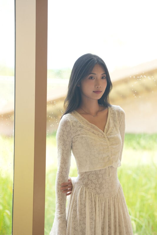 Una mujer con cabello largo y negro está parada en el interior, cerca de una ventana grande, con un vestido de encaje blanco. La suave luz natural se filtra a través de las ventanas y se puede ver la vegetación al fondo del exterior.