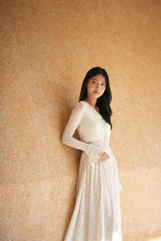 Una mujer que lleva un vestido largo de encaje blanco se encuentra junto a una pared texturizada de color marrón claro y mira a la cámara con una expresión amable. Una suave luz natural ilumina la escena.