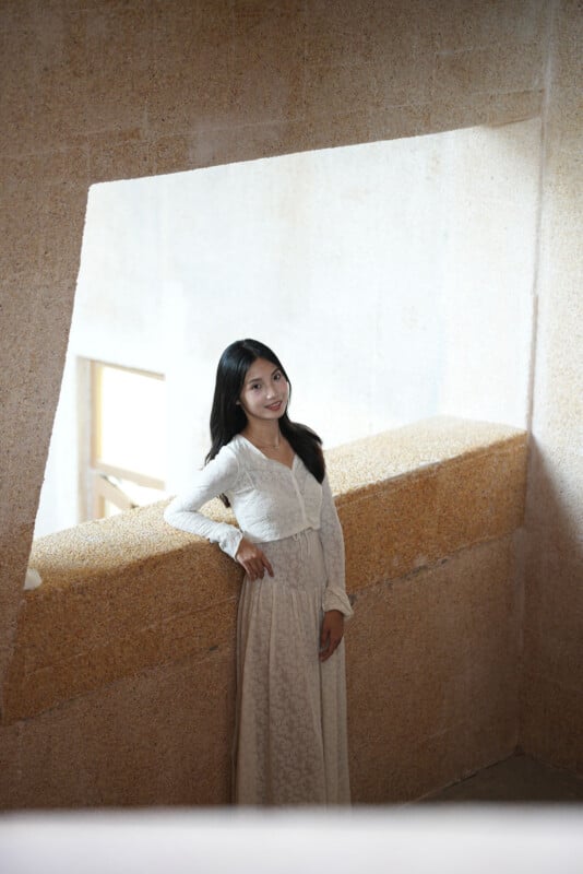 Una mujer con un vestido largo blanco se encuentra junto a una pared beige texturizada y sonríe suavemente a la cámara. La luz natural entra a través de las ventanas, creando un ambiente tranquilo y elegante.