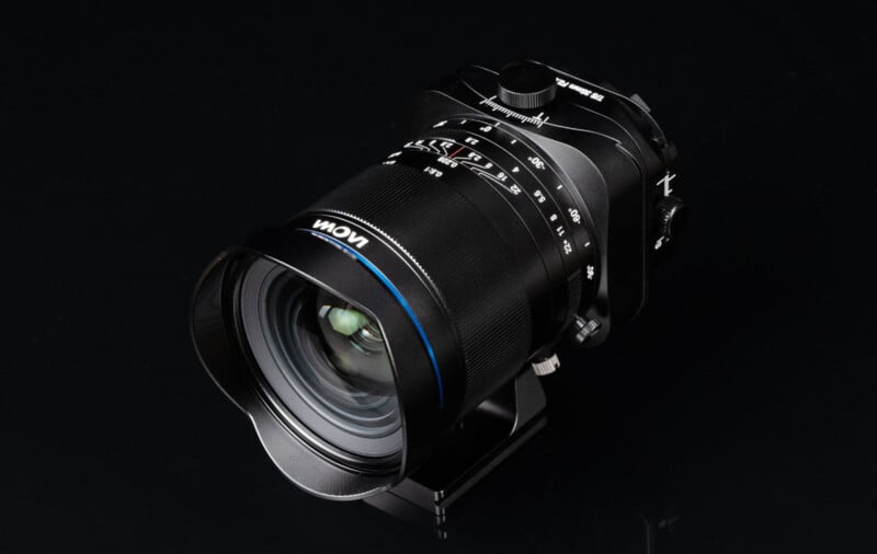 A close-up photo of a black Laowa camera lens with blue accents, mounted on a support bracket, against a black background. The lens displays various focus and aperture markings.