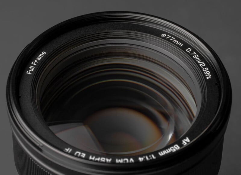 Primer plano de la lente de una cámara que muestra reflejos detallados en el cristal con marcas técnicas blancas que indican cosas como distancia focal de 85 mm, apertura de f/1,4, tamaño de filtro de 77 mm y "fotograma completo" compatibilidad.