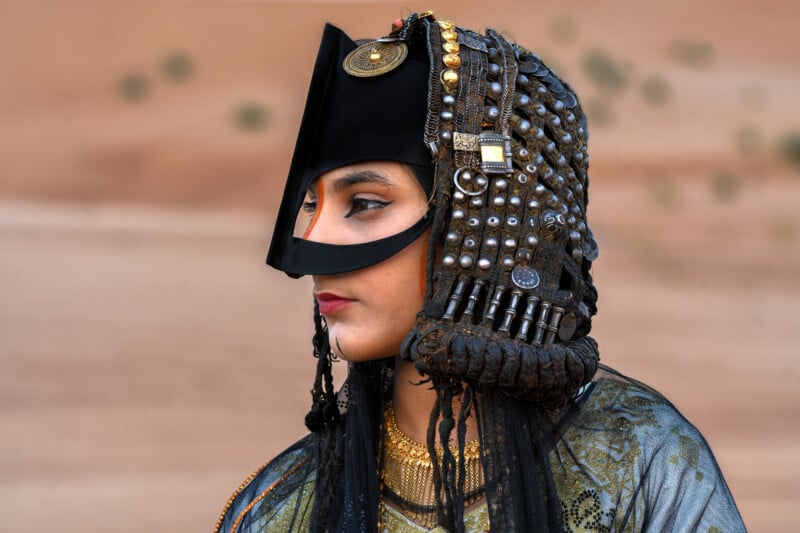 A woman in traditional attire wears an ornate headdress with metal adornments and a face covering, featuring intricate patterns and jewelry, against a blurred desert background.