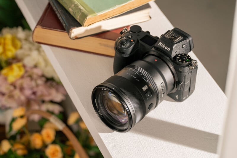 Cámara Nikon colocada sobre una superficie blanca junto a una pila de libros sobre un fondo de flores amarillas y rosadas suaves y desenfocadas.
