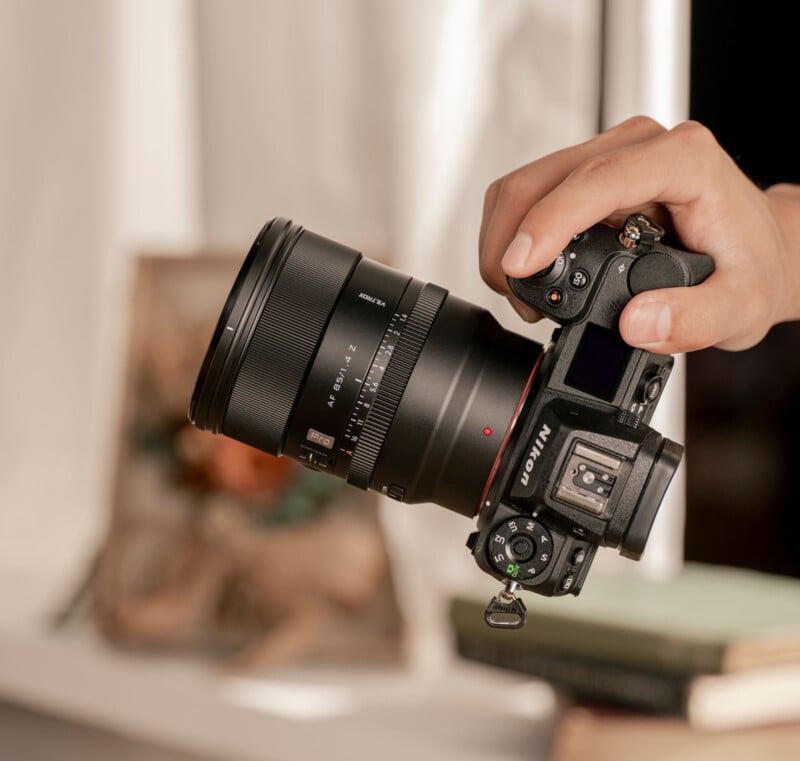 Una mano sostiene una cámara digital Nikon con una lente sigma grande, colocada a un lado cerca de la ventana. Al fondo se ven libros borrosos y objetos decorativos.