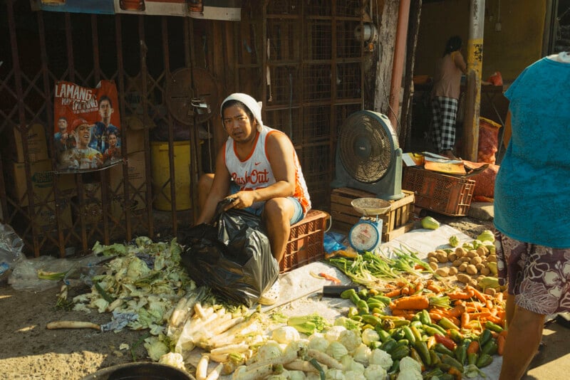 Un hombre con camisa sin mangas y turbante está sentado entre montones de verduras frescas en un puesto de mercado al aire libre, rodeado de zanahorias, coles y otros productos, con un ventilador y una balanza cerca.
