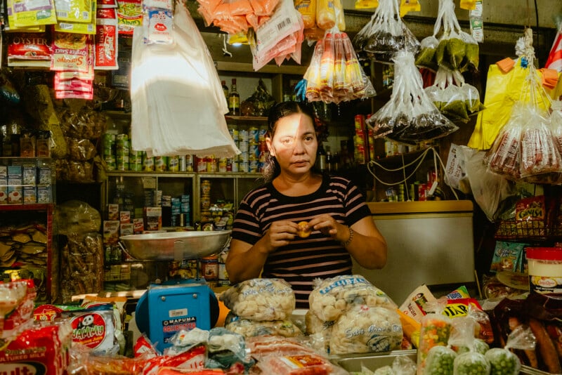 Una mujer se encuentra detrás de un puesto de mercado abarrotado de productos empaquetados, bocadillos y artículos colgantes. La luz del sol ilumina parcialmente su rostro mientras prepara la comida con las manos. Los productos en los estantes cubren el fondo.