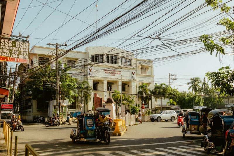 En un día soleado, la concurrida calle frente al edificio gubernamental se llena de triciclos, algunos coches, peatones y muchos cables eléctricos aéreos. Alrededor de la intersección se pueden ver palmeras y señales de tráfico.
