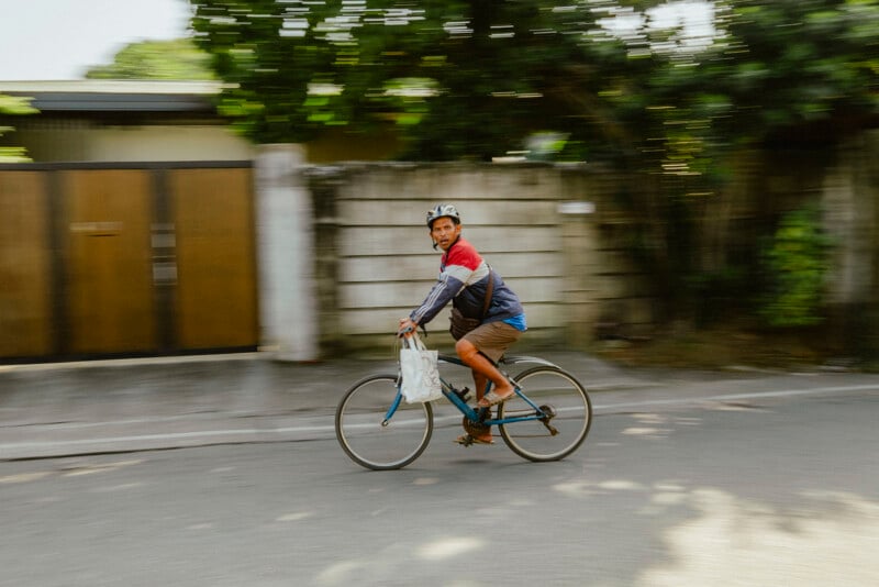 Un hombre con casco y chaqueta anda en bicicleta por la calle, con una bolsa colgando del manillar. El fondo es borroso, lo que sugiere movimiento, y se pueden ver árboles, paredes y una puerta marrón.