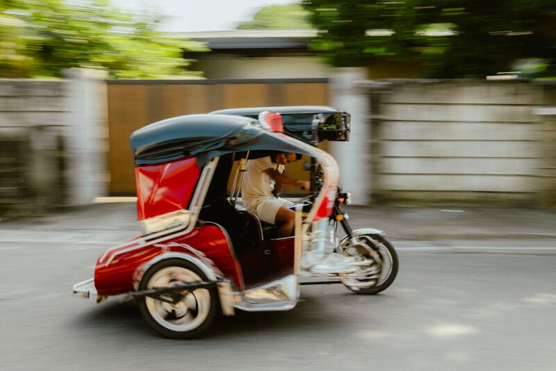 Un hombre conduce un triciclo motorizado rojo (comúnmente conocido como triciclo o tuk-tuk) en una calle de movimiento borroso con un muro de piedra y plantas verdes al fondo.