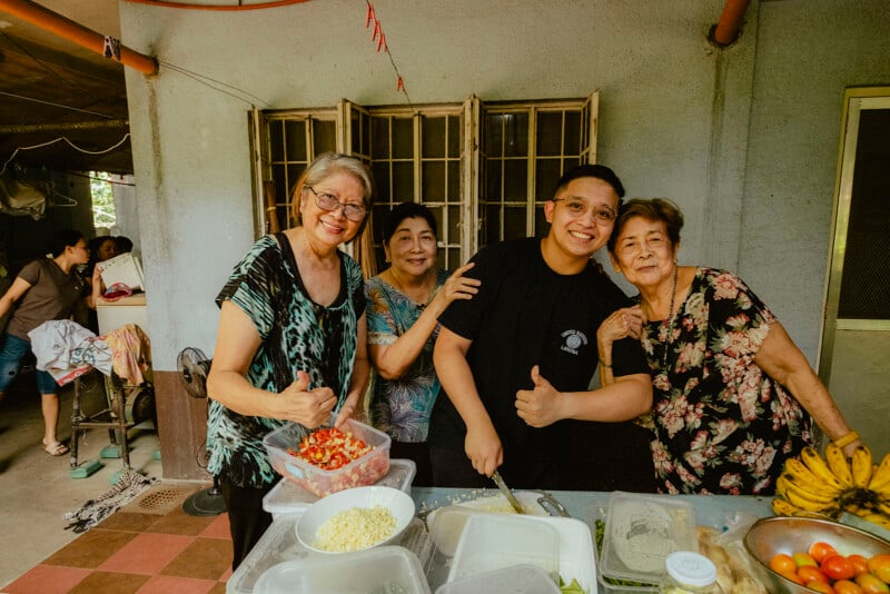 Cuatro personas sonrientes paradas alrededor de la mesa preparando comida y levantando el pulgar. Sobre la mesa se colocan verduras picadas, plátanos y otros ingredientes. El escenario está ambientado en una cocina familiar y el ambiente es relajado y alegre.