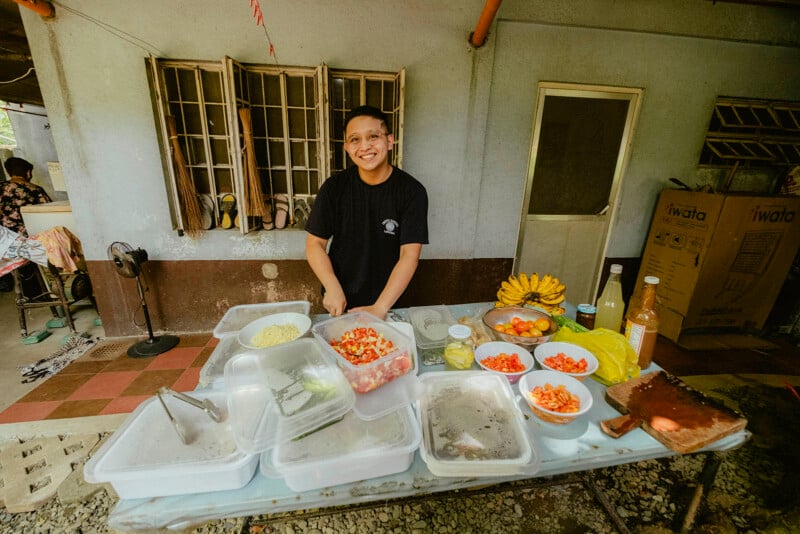 Un hombre sonriente prepara comida al aire libre, de pie detrás de una mesa llena de verduras picadas, plátanos, botellas y utensilios de cocina, y frente a una casa con las ventanas abiertas.