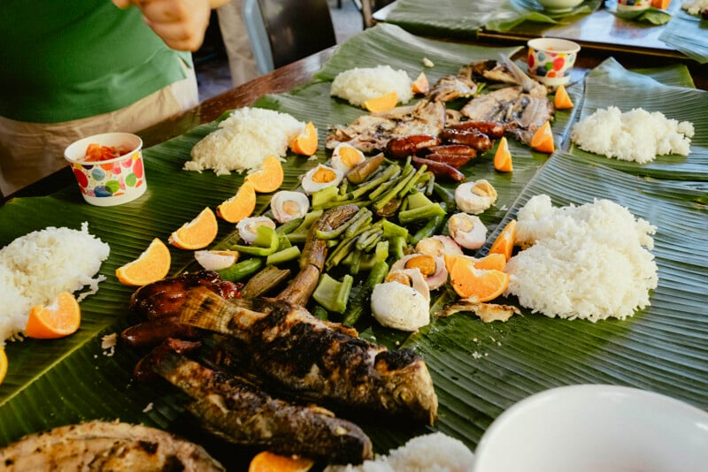 La tradicional comida de pelea filipina se sirve en una hoja de plátano e incluye pescado a la parrilla, arroz, vegetales verdes, salchichas y rodajas de naranja, con bebidas en vasos coloridos cerca.