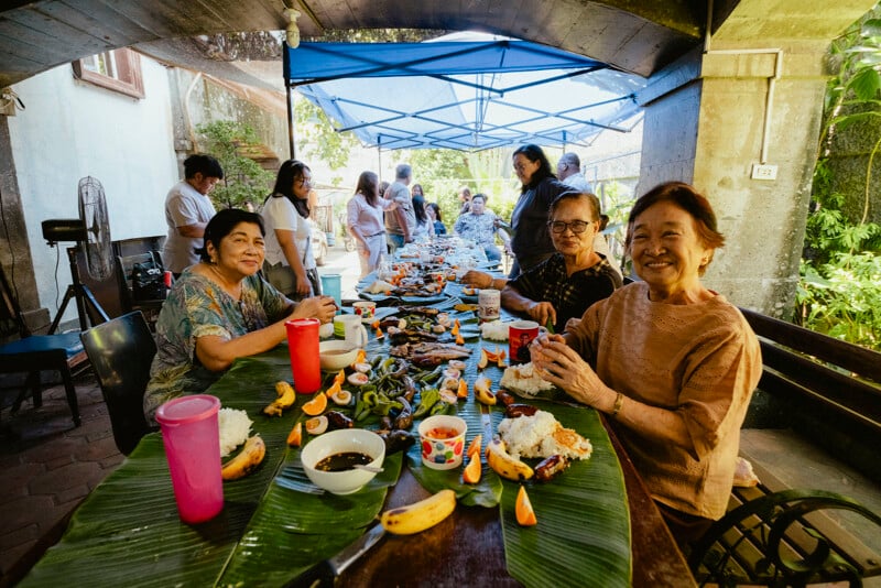 Un grupo de personas se reunió alrededor de una larga mesa cubierta con hojas de plátano para compartir una comida al aire libre. Delante estaban sentados tres ancianos, sonrientes, con platos de comida y fruta delante. Otros hablan de fondo.