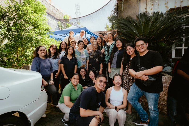 Un grupo de personas sonriendo y posando juntas en un jardín soleado con plantas verdes, un auto blanco y una casa al fondo. Parecen estar celebrando o reuniéndose para una ocasión especial.