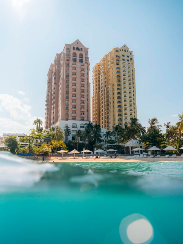 Dos altos y coloridos edificios frente al mar se elevan por encima de las palmeras y sombrillas de la playa, visibles desde el agua turquesa con la luz del sol reflejándose en el agua.